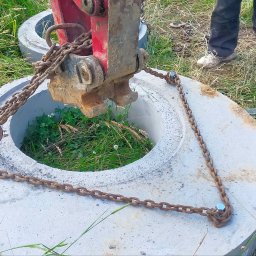 Zbigniew Kańtoch - Betonowy krąg studzienny podnoszony przez maszynę z użyciem łańcuchów, widoczna trawa wewnątrz i wokół kręgu, częściowo widoczny operator i drugi krąg w tle.