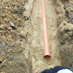 Zbigniew Kańtoch - Wąski wykop w piaszczystej ziemi z ułożoną rurą PCV, widoczni pracownicy i poziomica w tle.