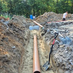 Zbigniew Kańtoch - Głęboki wąski wykop z ułożoną rurą kanalizacyjną z tworzywa sztucznego, obok łopata, w tle betonowy krąg studzienny i przenośna toaleta, ziemia usypana po bokach wykopu, w oddali drzewa.