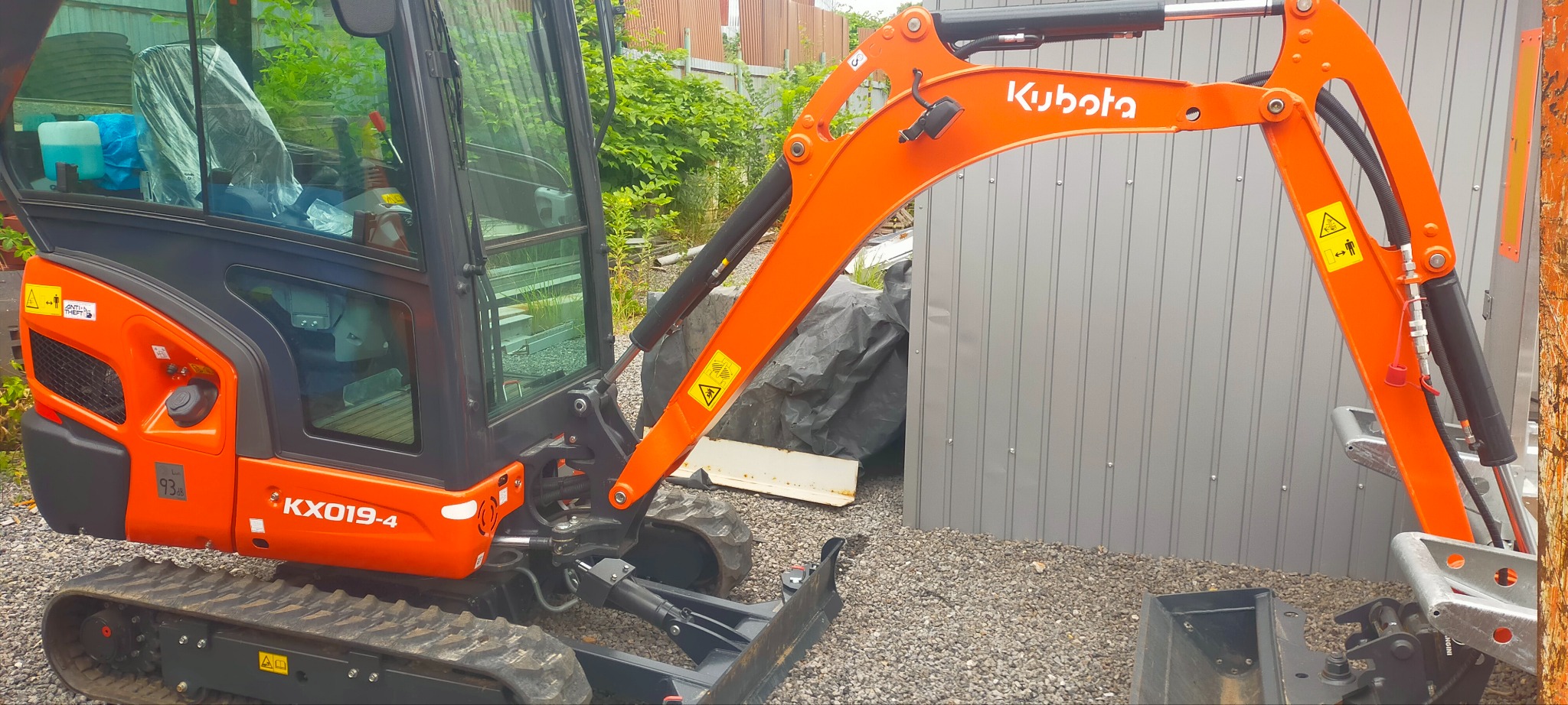 Pomarańczowa minikoparka Kubota KX019-4 na żwirowym podłożu, z widocznym ramieniem i łyżką, zaparkowana obok metalowego garażu i zieleni.