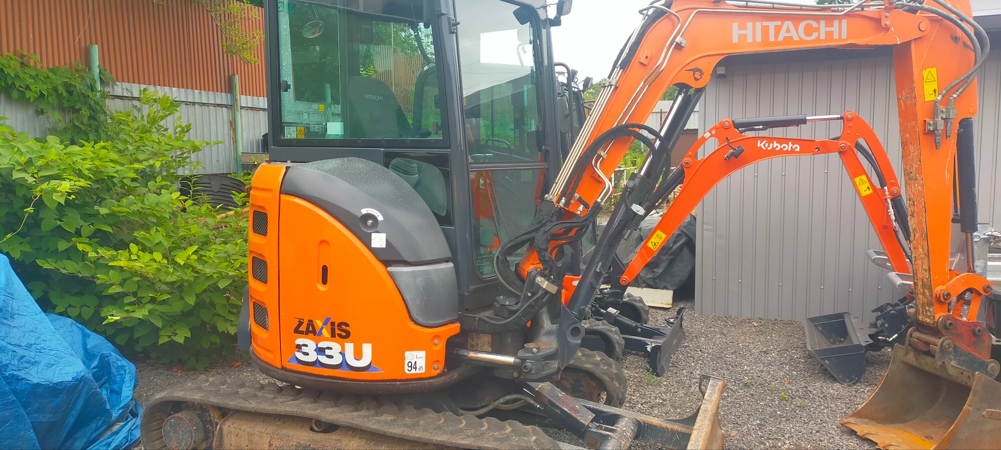 Pomarańczowa minikoparka Hitachi Zaxis 33U zaparkowana na żwirowym podłożu, z widoczną łyżką i ramieniem, na tle metalowego garażu i zieleni.