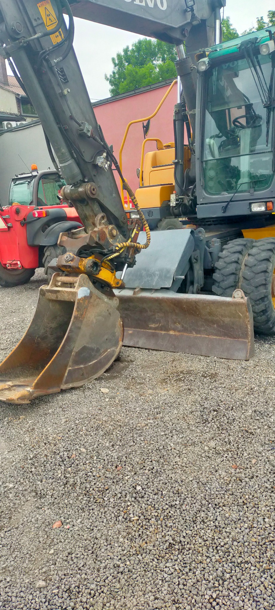 Żółto-szara koparka Volvo z łyżką i lemieszem na żwirowym podłożu, w tle czerwony podnośnik teleskopowy i budynek.