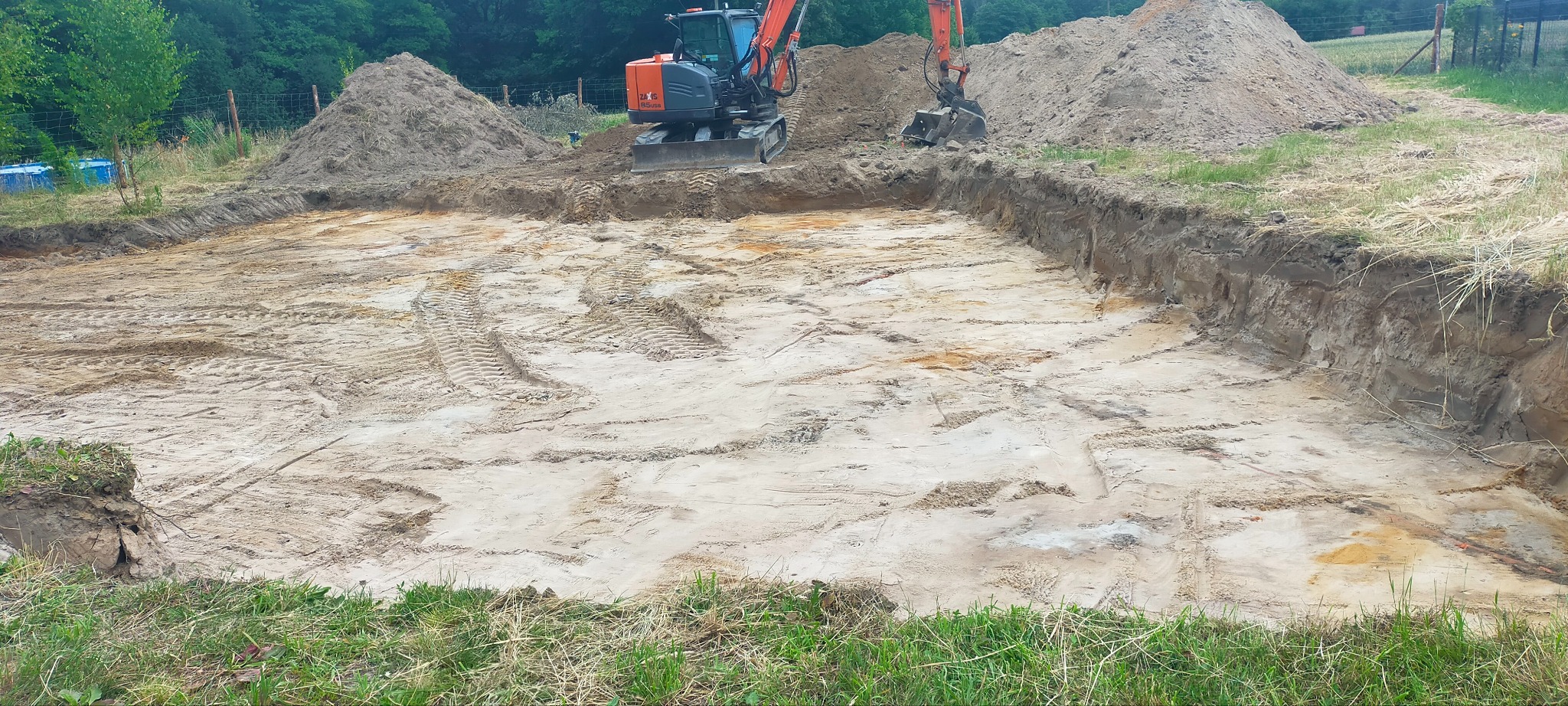 Pomarańczowa koparka Hitachi Zaxis 16 na placu budowy podczas wykopu prostokątnego dołu, widoczne ślady opon i pryzmy ziemi.