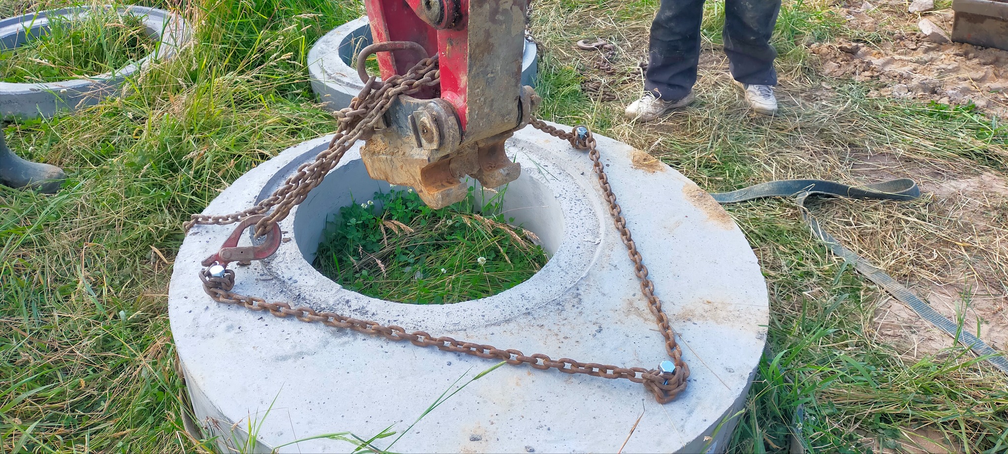 Betonowy krąg studzienny podnoszony przez maszynę z użyciem łańcuchów, widoczna trawa wewnątrz i wokół kręgu, częściowo widoczny operator i drugi krąg w tle.