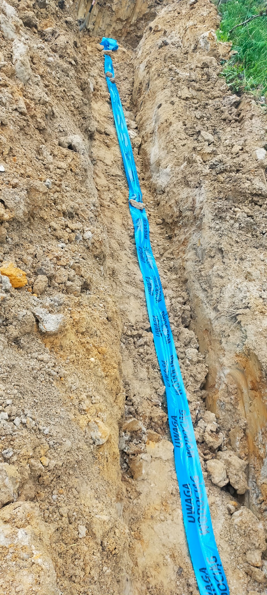 Wykop w ziemi z niebieską taśmą ostrzegawczą 'Uwaga' ułożoną wzdłuż wykopu, prawdopodobnie zabezpieczającą ułożone w nim instalacje.