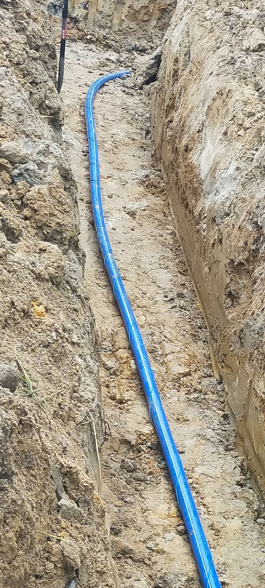 Niebieska rura w wąskim wykopie ziemnym, zabezpieczona niebieskim sznurkiem, widok z góry na piaszczyste dno i nierówne ściany wykopu.
