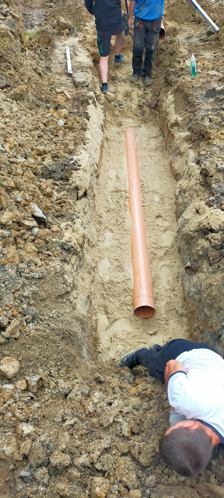 Wąski wykop w piaszczystej ziemi z ułożoną rurą PCV, widoczni pracownicy i poziomica w tle.