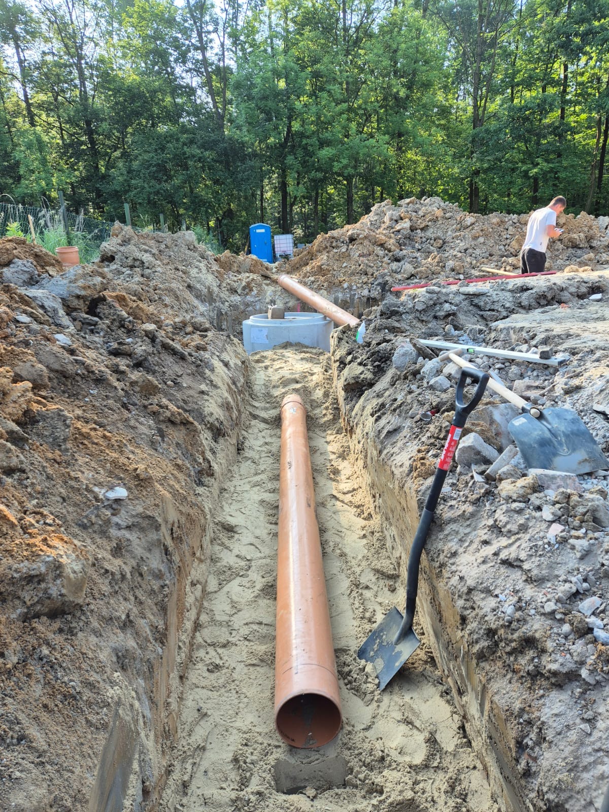 Głęboki wąski wykop z ułożoną rurą kanalizacyjną z tworzywa sztucznego, obok łopata, w tle betonowy krąg studzienny i przenośna toaleta, ziemia usypana po bokach wykopu, w oddali drzewa.