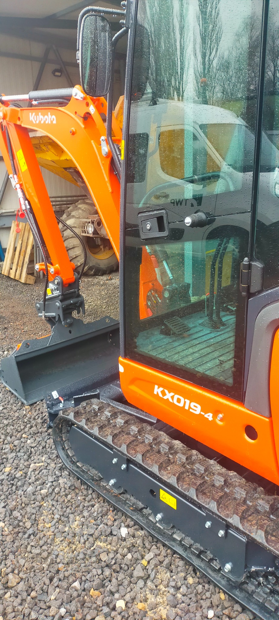 Pomarańczowa minikoparka Kubota KX019-4 z łyżką, stojąca na żwirowym podłożu, z widoczną kabiną i gąsienicami, z odbiciem białego samochodu dostawczego w szybie.
