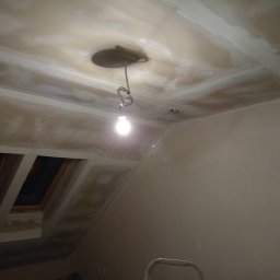 Dawid Mendyk - Poddasze w trakcie prac wykończeniowych: sufit z płyt kartonowo-gipsowych przygotowany do szpachlowania, widoczne łączenia płyt, wisząca żarówka i okna dachowe.