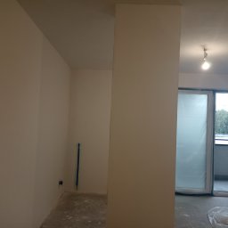 Dawid Mendyk - Wnętrze w trakcie prac wykończeniowych: świeżo pomalowane na jasny kolor ściany, zabezpieczone folią drzwi balkonowe, widoczna instalacja elektryczna i worek z zaprawą na podłodze.