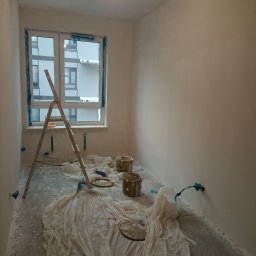 Dawid Mendyk - Pokój w trakcie prac wykończeniowych w Warszawie: ściany po nałożeniu gładzi, zabezpieczone folią podłoga i okno, drabina oparta o ścianę, widoczne puszki z masą szpachlową.