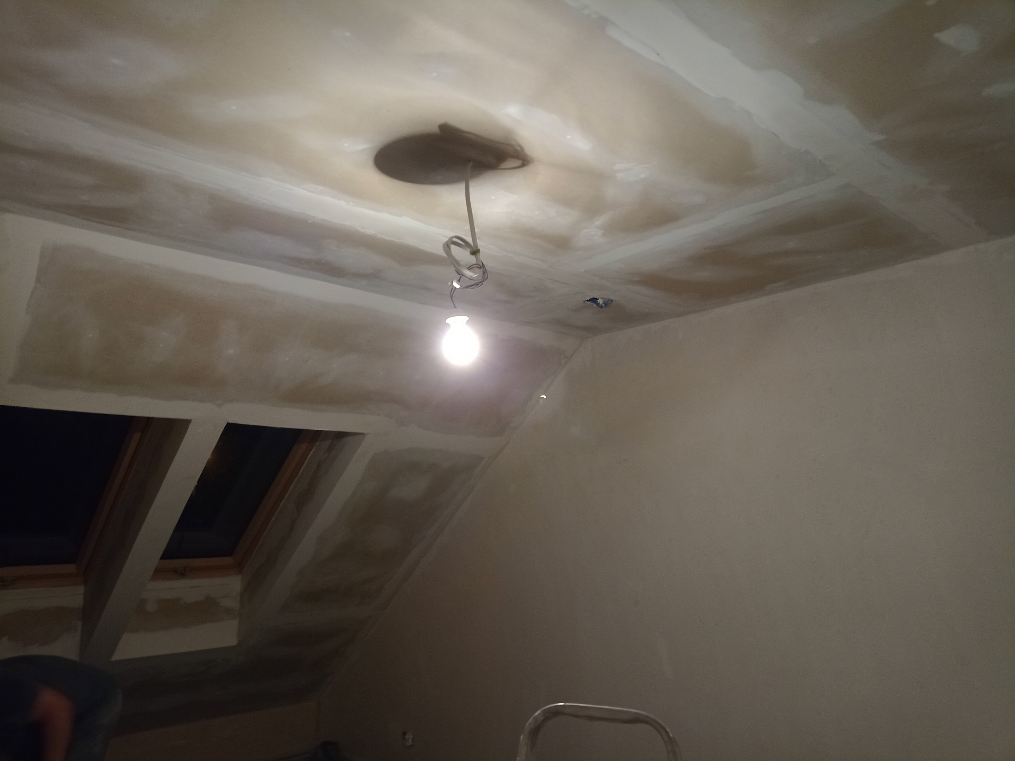 Poddasze w trakcie prac wykończeniowych: sufit z płyt kartonowo-gipsowych przygotowany do szpachlowania, widoczne łączenia płyt, wisząca żarówka i okna dachowe.