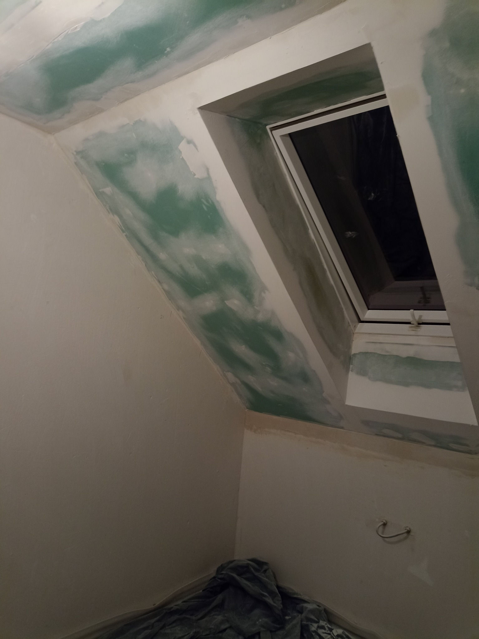 Poddasze z oknem dachowym w trakcie szpachlowania, widoczne ślady po nałożonej masie szpachlowej na ścianach i suficie.