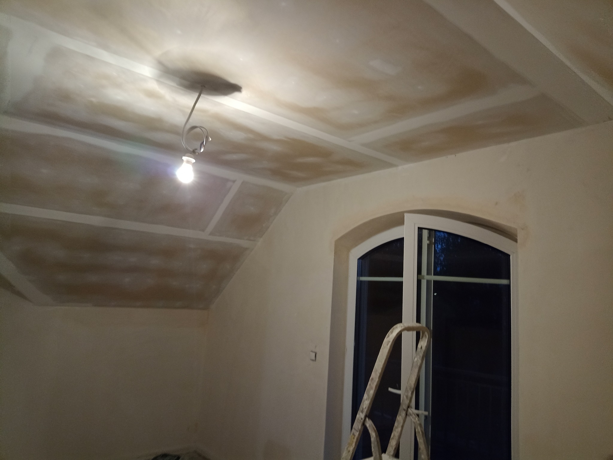 Pokój w trakcie prac przygotowawczych: sufit z płyt kartonowo-gipsowych przed szpachlowaniem, widoczne łączenia płyt, wisząca żarówka i drabina oparta o okno.