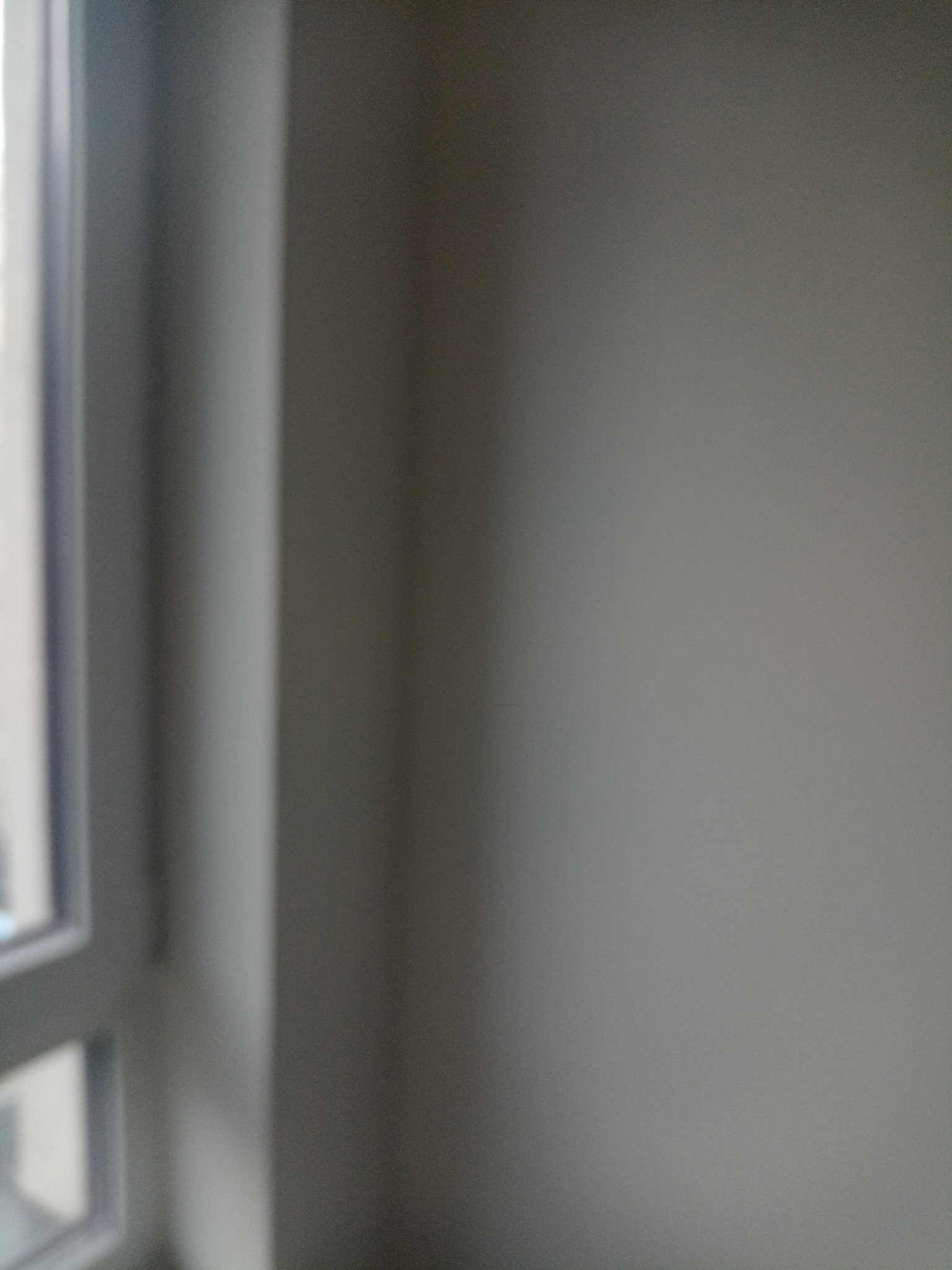 Nierównomierne oświetlenie szarej ściany z fragmentem ramy okiennej po lewej stronie, z widocznymi refleksami światła.