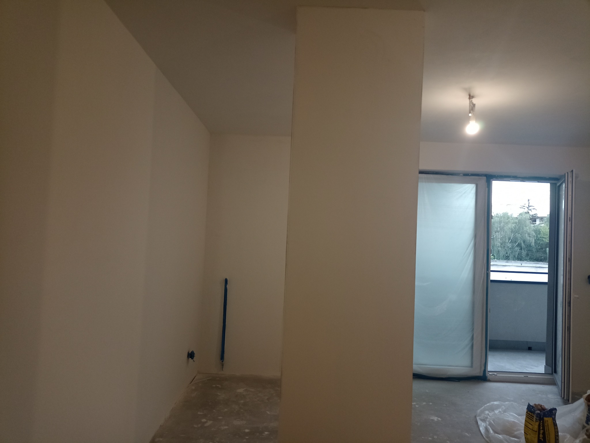 Wnętrze w trakcie prac wykończeniowych: świeżo pomalowane na jasny kolor ściany, zabezpieczone folią drzwi balkonowe, widoczna instalacja elektryczna i worek z zaprawą na podłodze.