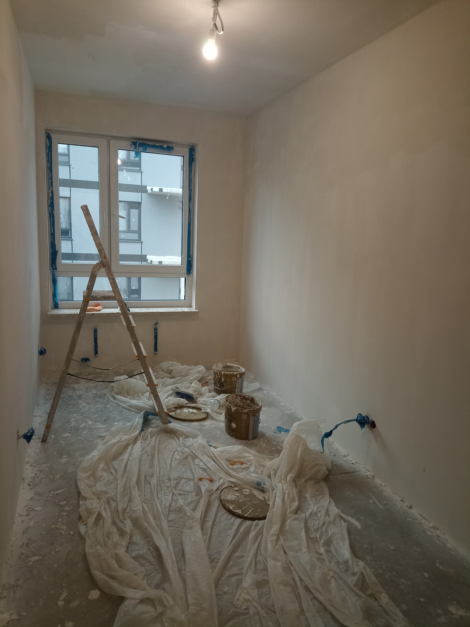 Pokój w trakcie prac wykończeniowych w Warszawie: ściany po nałożeniu gładzi, zabezpieczone folią podłoga i okno, drabina oparta o ścianę, widoczne puszki z masą szpachlową.