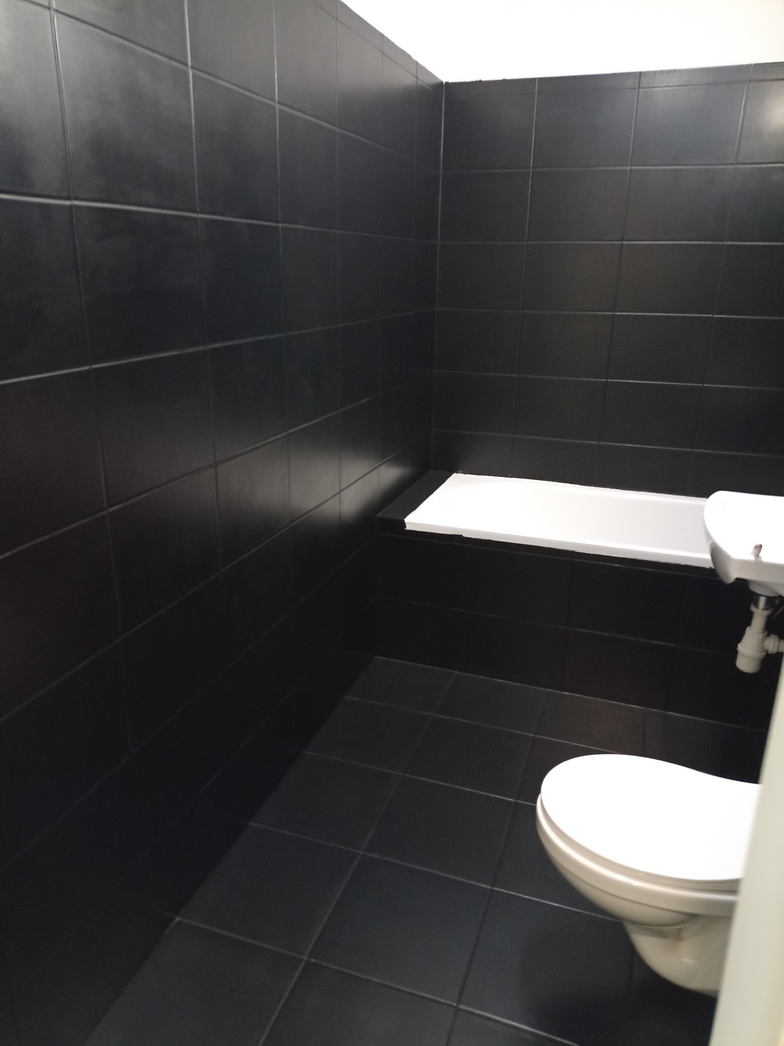 Minimalistyczna łazienka z czarnymi płytkami na ścianach i podłodze, biała umywalka i toaleta, surowy wystrój.