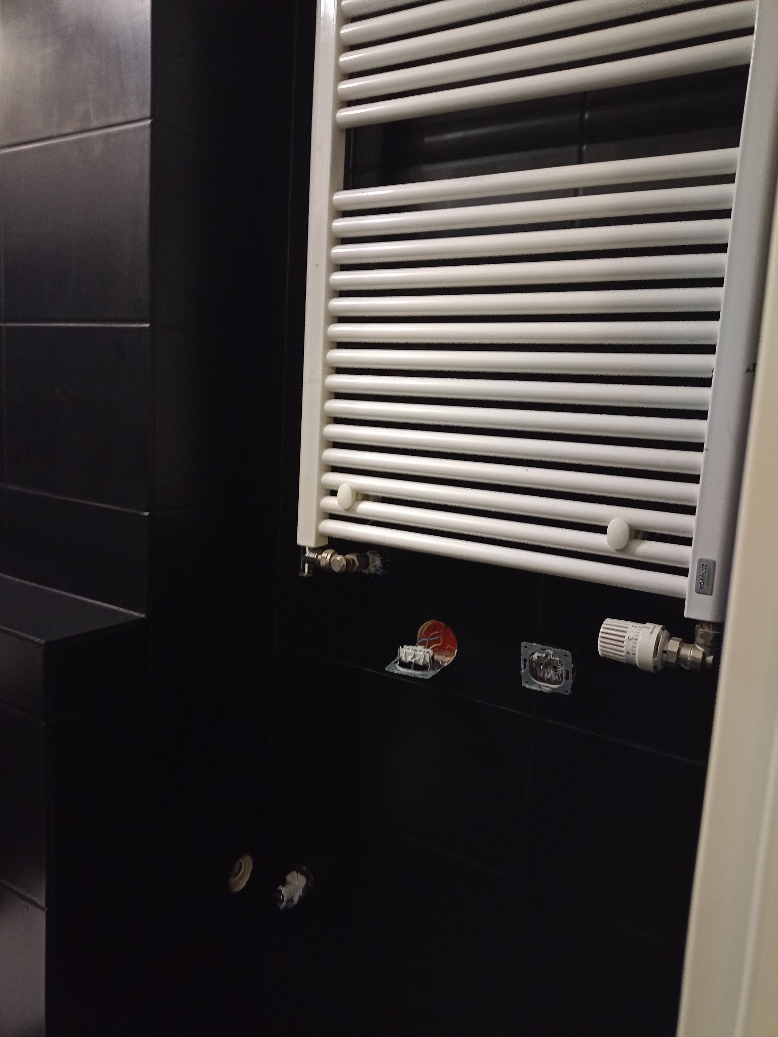 Biały grzejnik drabinkowy zamontowany na czarnej ścianie z płytek, widoczne niedokończone podłączenia hydrauliczne i elektryczne, w tle fragment ściany