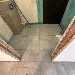 valentyn haliev - Łazienka w trakcie remontu: ułożone szare płytki podłogowe, częściowo wykończone ściany z widocznymi instalacjami, poziomica oparta o ścianę, widoczny fragment buta osoby robiącej zdjęcie.