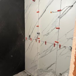 valentyn haliev - Ściana łazienki w trakcie układania płytek imitujących marmur w kolorze białym i czarnym; widoczne czerwone klipsy poziomujące, przewody elektryczne i drabinka.