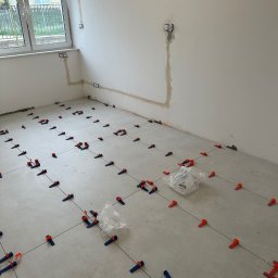 valentyn haliev - Podłoga w trakcie układania płytek, z systemem poziomowania składającym się z klipsów i klinów w kolorach niebieskim i pomarańczowym, widoczne krzyżyki dystansowe i żyłka, ściany w stanie surowym...