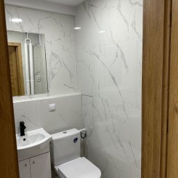 valentyn haliev - Wykończona łazienka z białymi płytkami imitującymi marmur, białą szafką z umywalką i czarną armaturą, oraz toaletą. Widoczne drewniane drzwi i lustro.