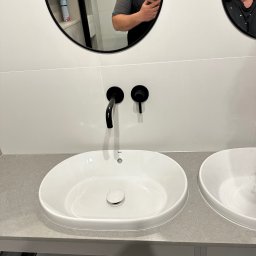 valentyn haliev - Dwie białe umywalki nablatowe z czarną armaturą zamontowane na szarym blacie, widoczne elementy montażowe szafki podumywalkowej oraz odbicie dwóch osób w lustrach nad umywalkami.