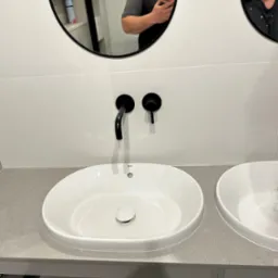 Dwie białe umywalki nablatowe z czarną armaturą zamontowane na szarym blacie, widoczne elementy montażowe szafki podumywalkowej oraz odbicie dwóch osób w lustrach nad umywalkami.