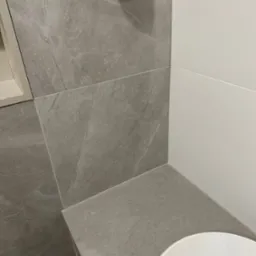 Fragment wykończonej łazienki z szarymi płytkami imitującymi marmur na ścianach i blacie, połączonymi z białymi panelami ściennymi, widoczny fragment umywalki.