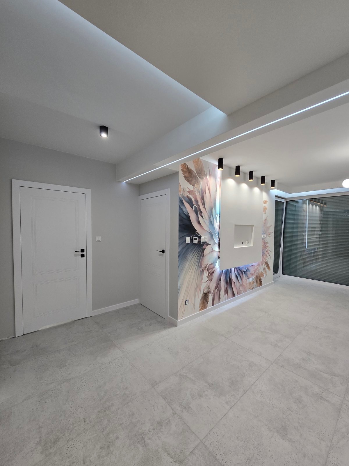 Nowoczesne wnętrze z szarą podłogą, białymi drzwiami i ścianą z abstrakcyjnym motywem kwiatowym, oświetlenie sufitowe LED i czarne reflektory.