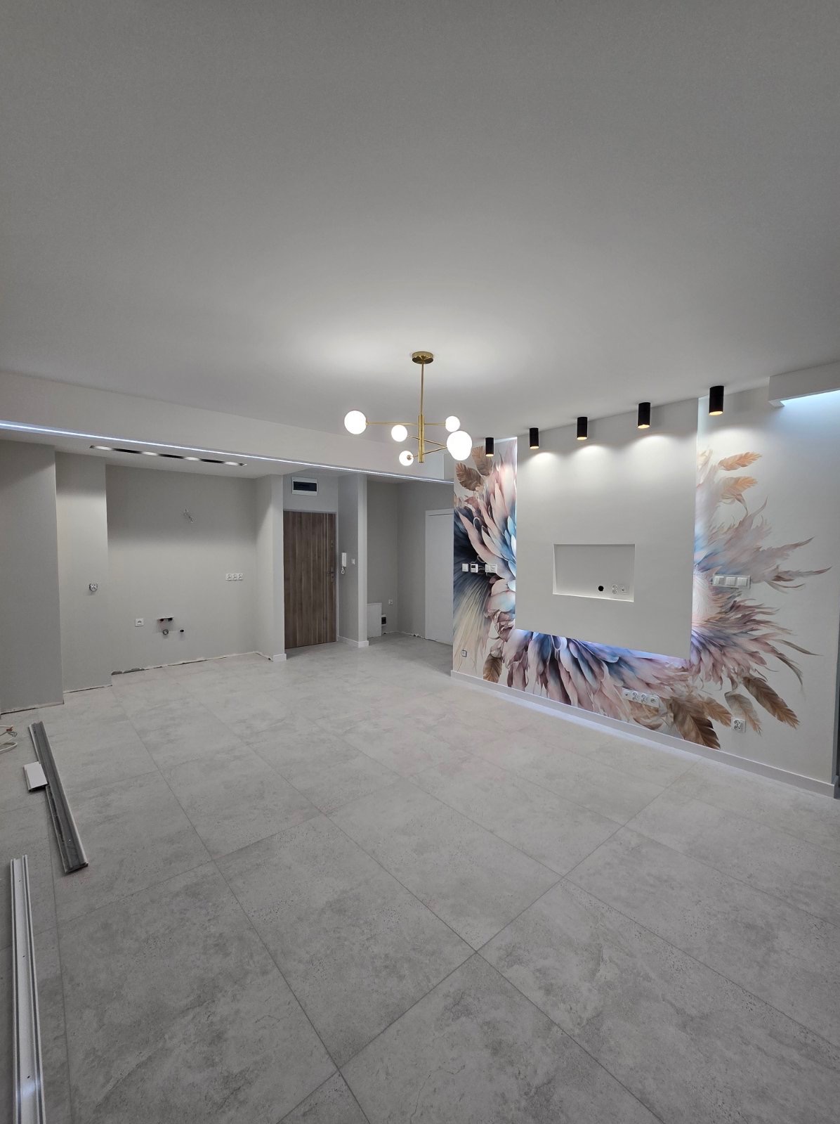 Wnętrze pokoju z szarą podłogą z płytek, ozdobną tapetą z motywem piór na ścianie, nowoczesnym oświetleniem sufitowym i listwami przypodłogowymi w trakcie montażu.