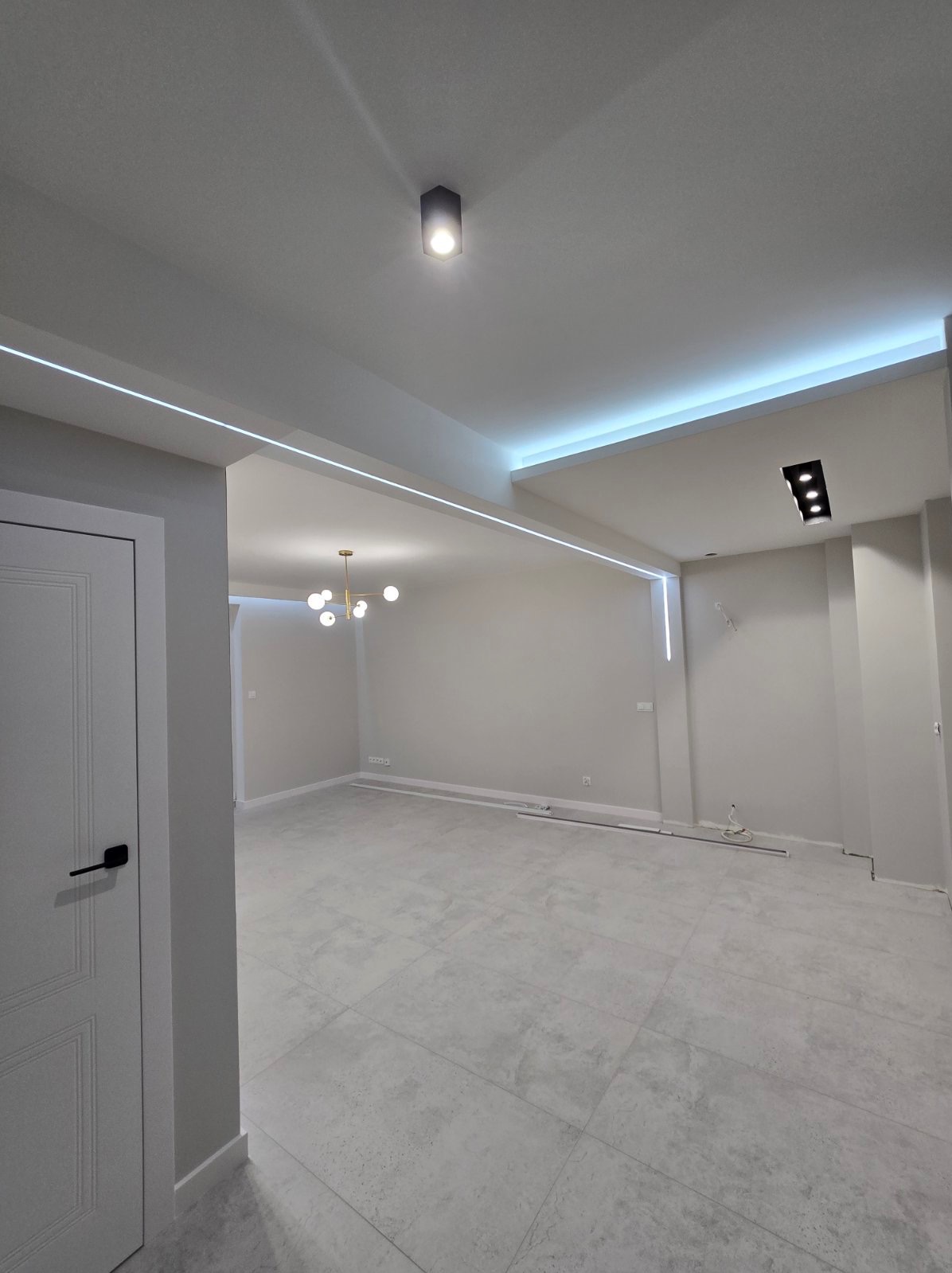 Szare wnętrze po remoncie z podświetleniem LED na suficie, widoczne płytki podłogowe i nowoczesna lampa wisząca w oddali.