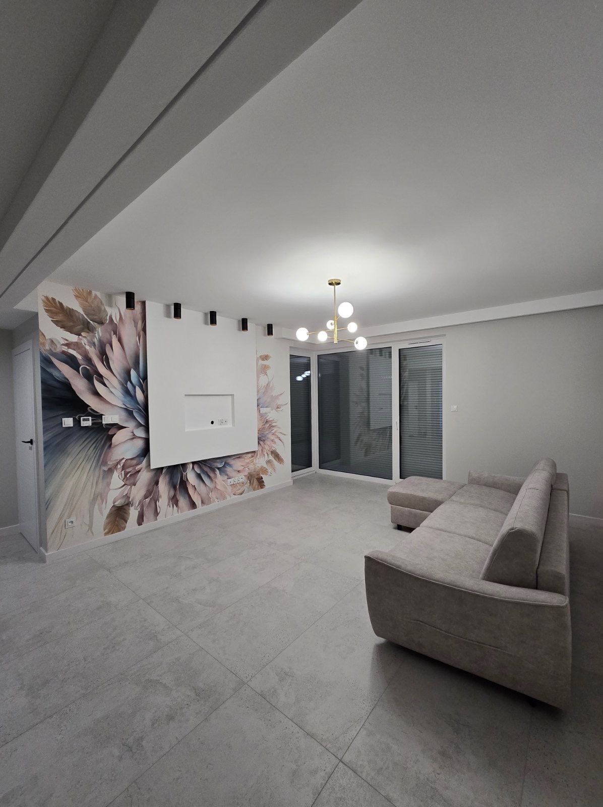 Salon w jasnych barwach z szarą sofą narożną, ścianą z motywem kwiatowym i nowoczesnym oświetleniem sufitowym.