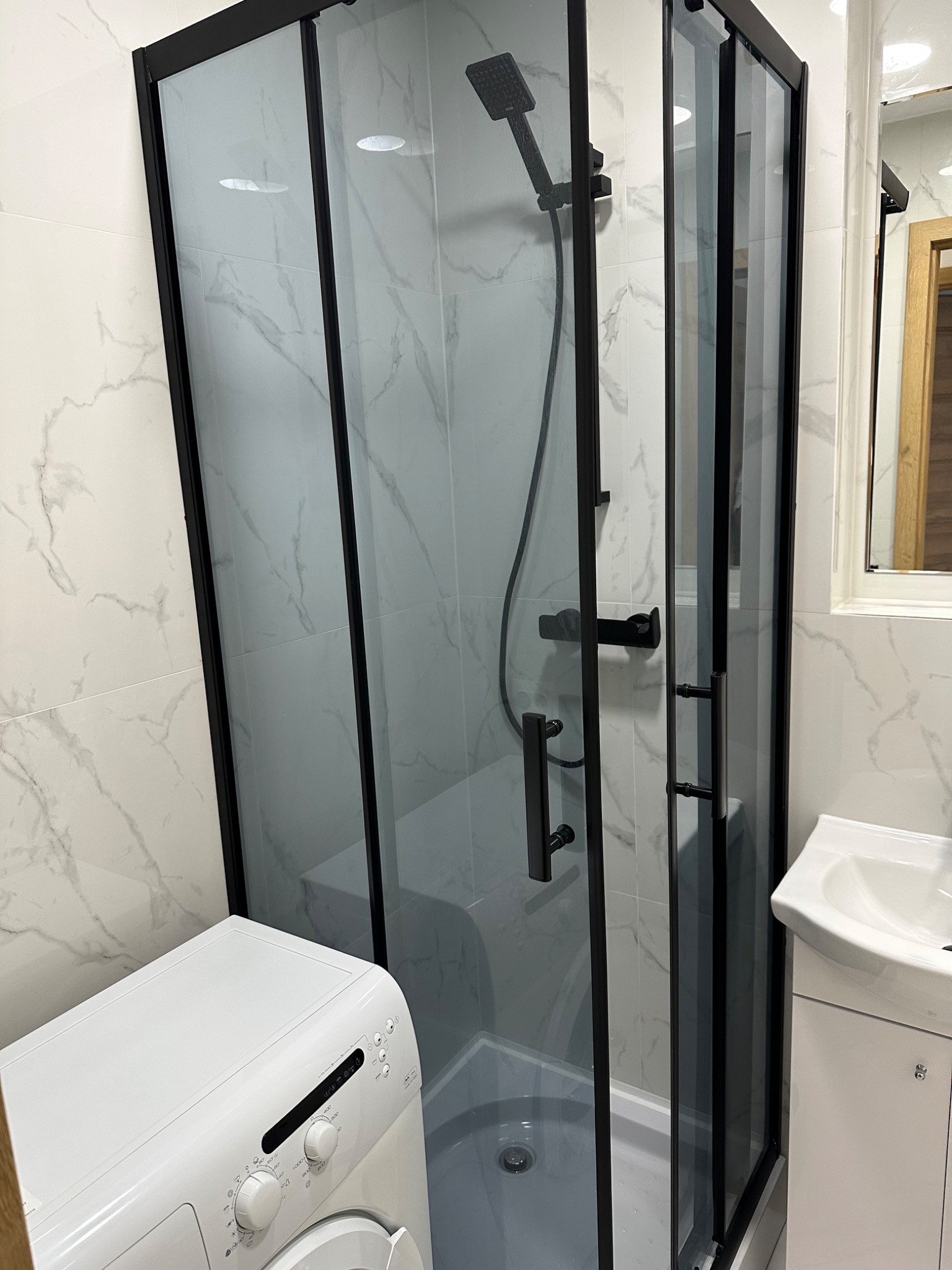 Nowoczesna kabina prysznicowa z czarnymi profilami i deszczownicą, białe marmurowe płytki na ścianach, pralka i umywalka w tle.