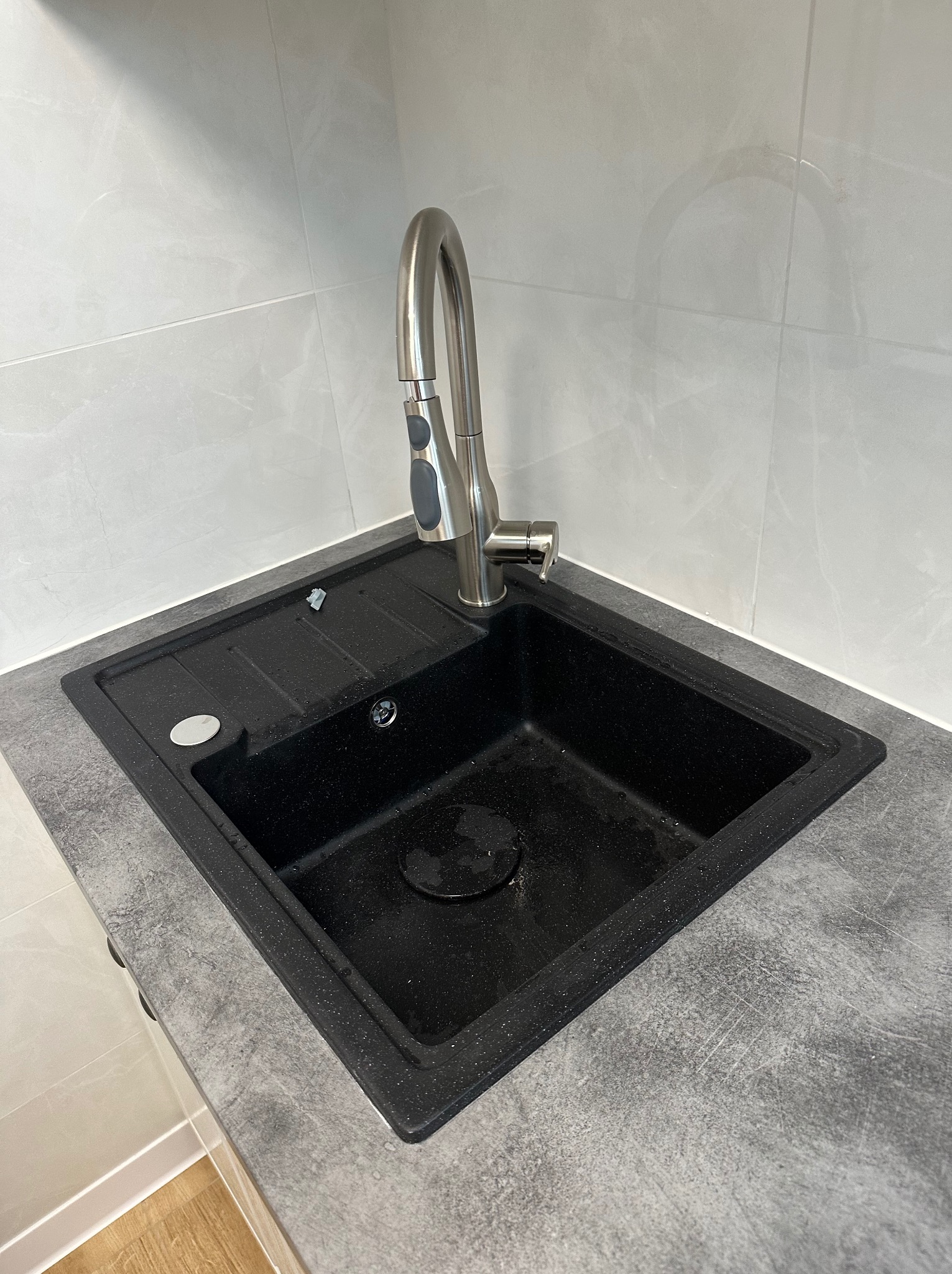 Czarny, granitowy zlew kuchenny z chromowaną, wyciąganą wylewką, osadzony w blacie o teksturze betonu, na tle jasnych, marmurkowych płytek ściennych.
