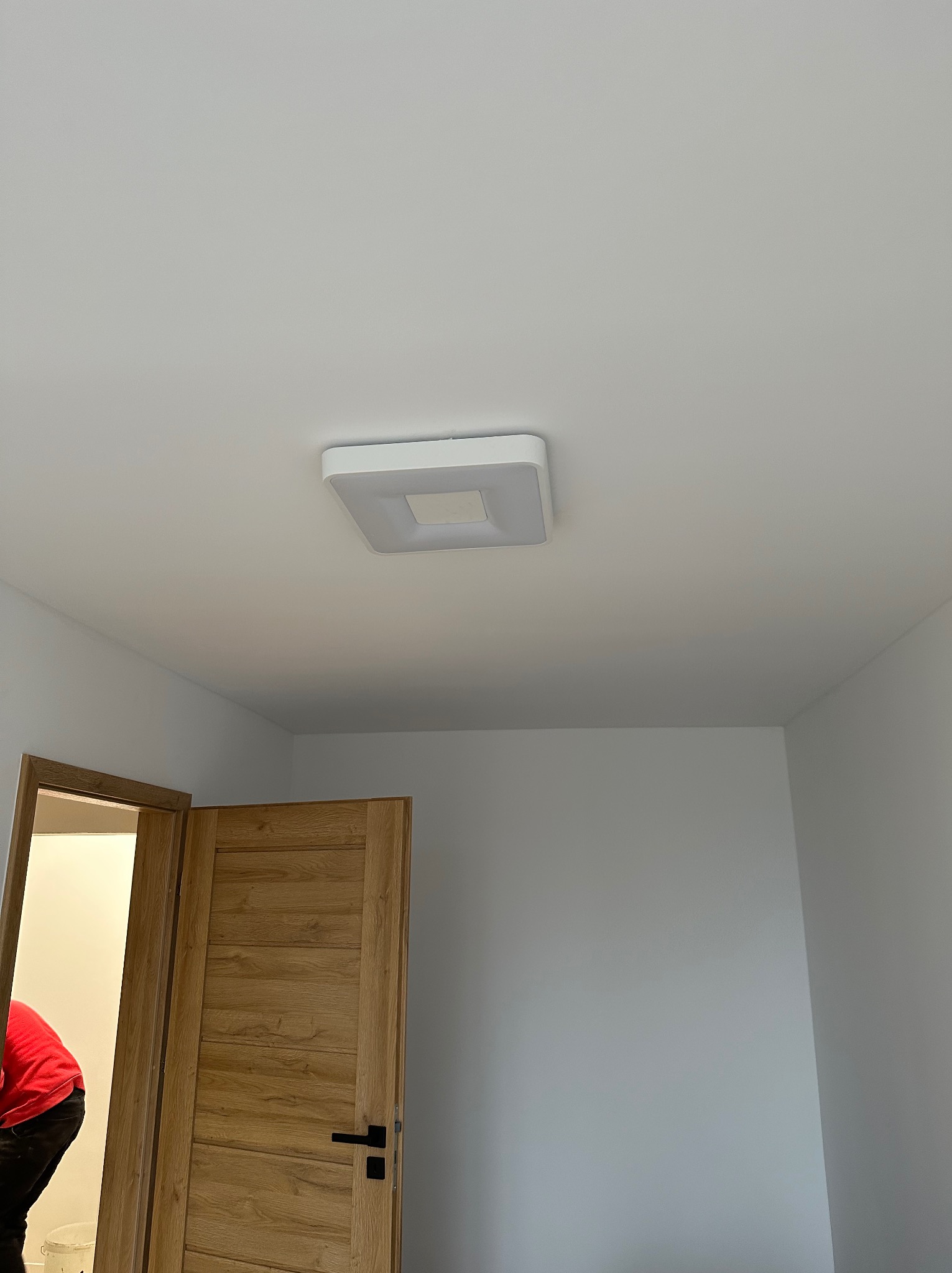 Białe wnętrze z zamontowaną kwadratową lampą sufitową LED i otwartymi drewnianymi drzwiami z czarną klamką, widoczny fragment osoby w czerwonej koszulce.