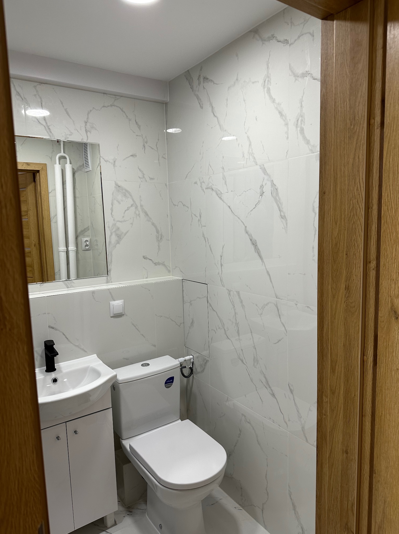 Wykończona łazienka z białymi płytkami imitującymi marmur, białą szafką z umywalką i czarną armaturą, oraz toaletą. Widoczne drewniane drzwi i lustro.