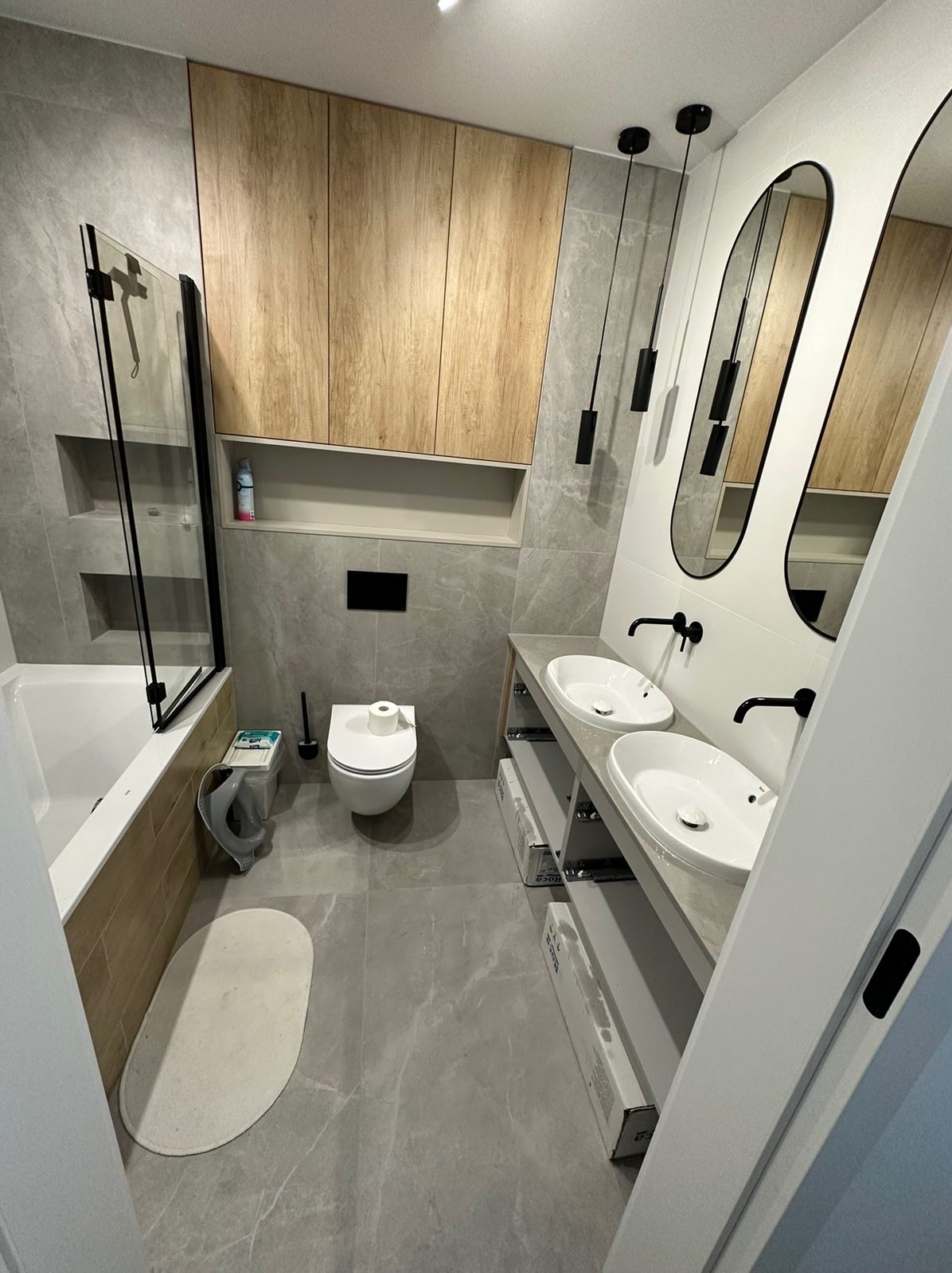 Nowoczesna łazienka z szarymi płytkami imitującymi marmur, drewnianymi szafkami wiszącymi, dwoma owalnymi umywalkami nablatowymi z czarną armaturą, podłużnymi lustrami w czarnych ramach i wiszącymi...