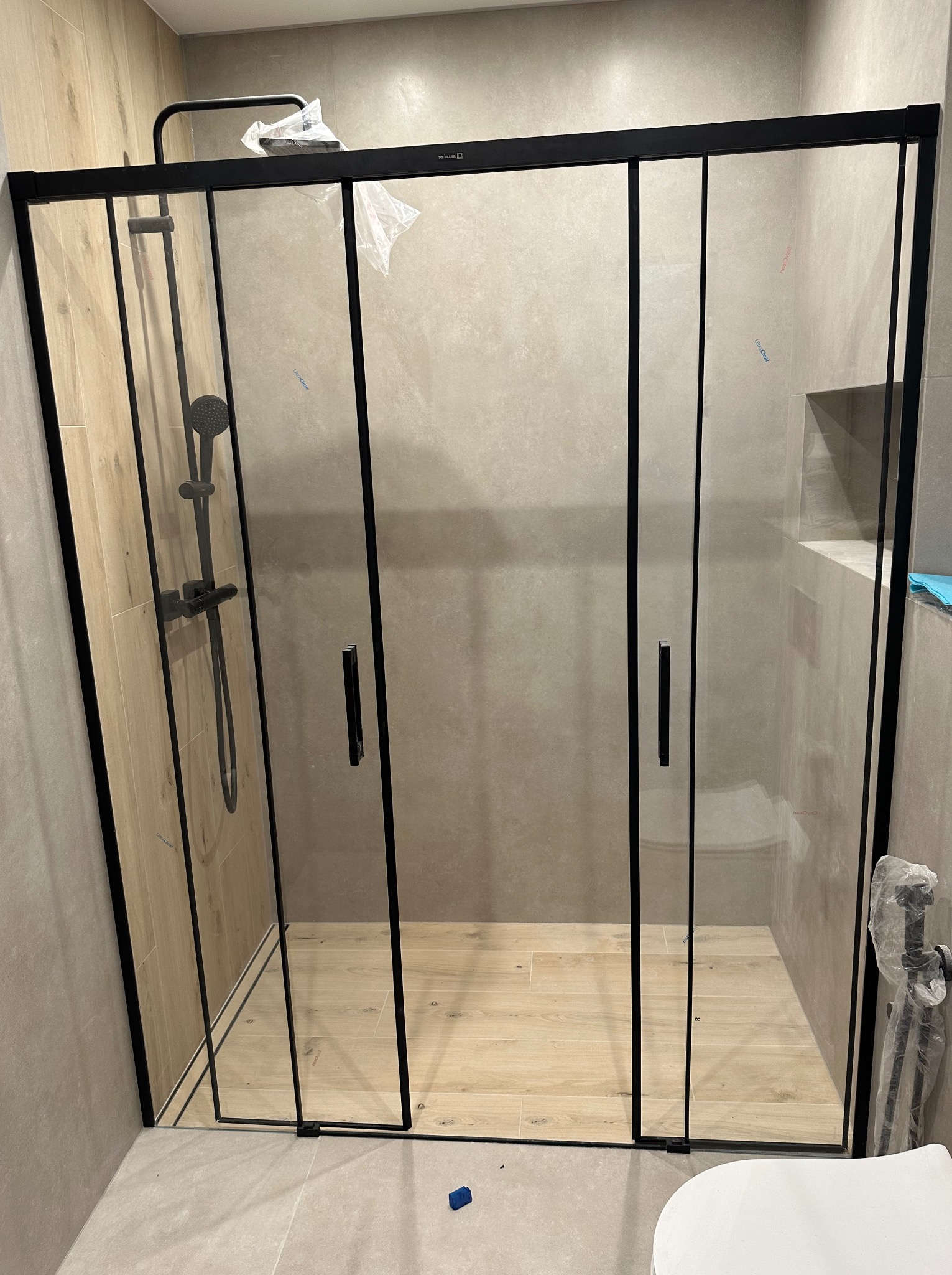 Nowoczesna kabina prysznicowa z czarnymi profilami i szklanymi drzwiami przesuwnymi, zamontowana w łazience wykończonej płytkami w odcieniach beżu i szarości, widoczna bateria prysznicowa w kolorze...