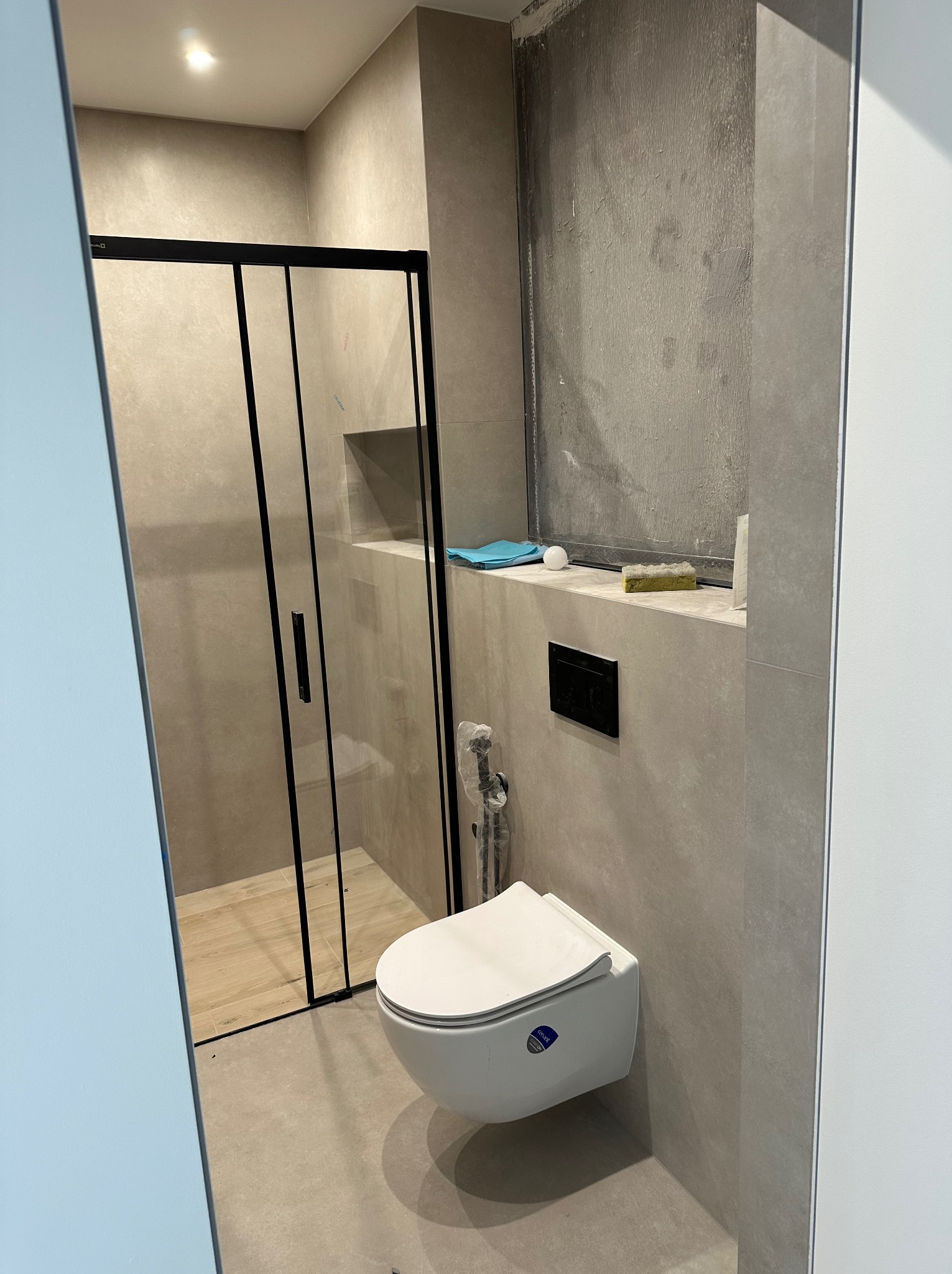 Wnętrze łazienki w trakcie prac wykończeniowych; widoczna kabina prysznicowa z czarnymi profilami, podwieszana miska WC, fragment ściany przygotowanej pod położenie płytek, niebieska szmatka...