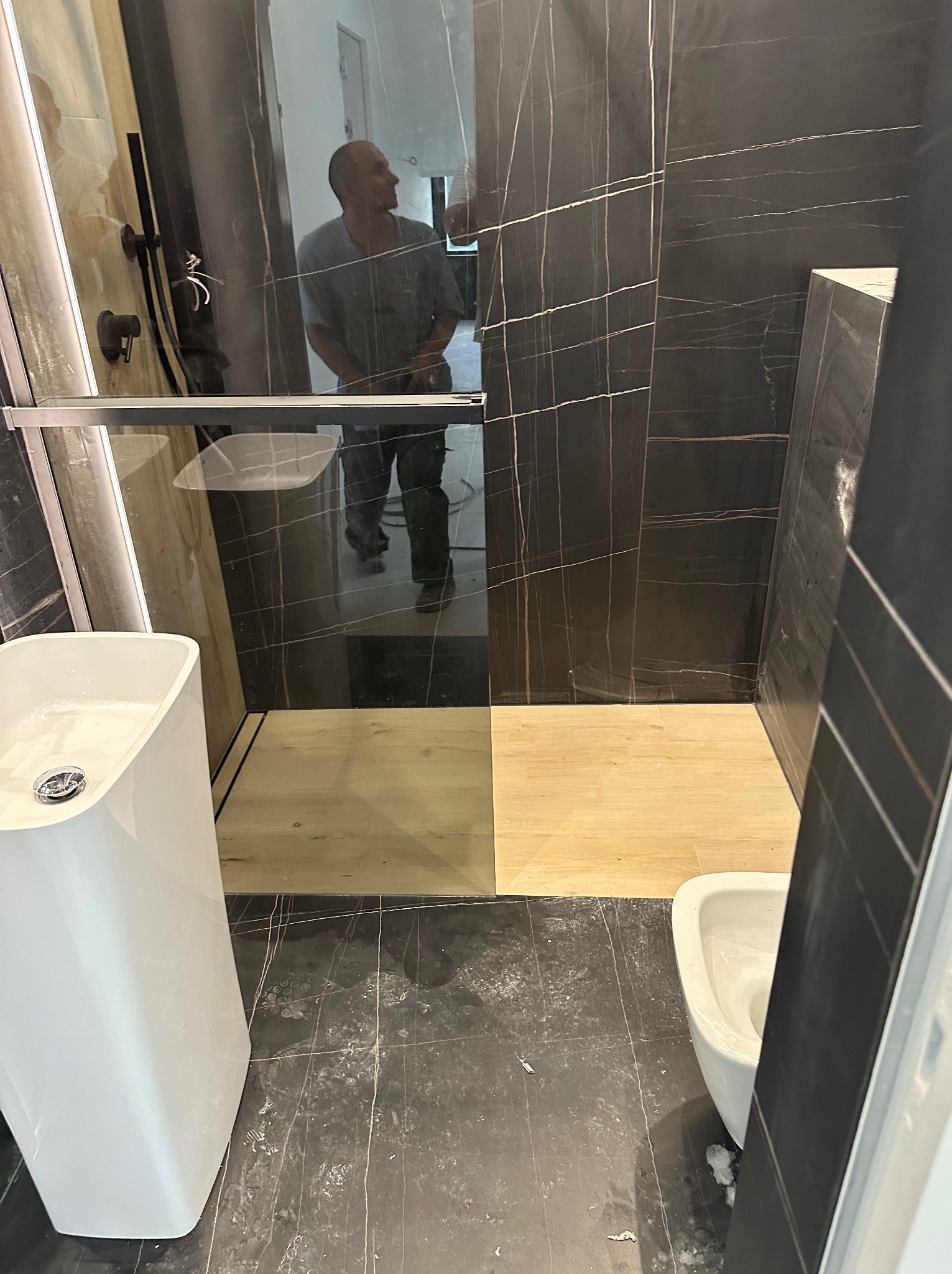 Nowoczesna łazienka z czarnymi płytkami imitującymi marmur, kabiną prysznicową typu walk-in z drewnianym brodzikiem, białą umywalką nablatową i toaletą w trakcie prac wykończeniowych, widoczny...