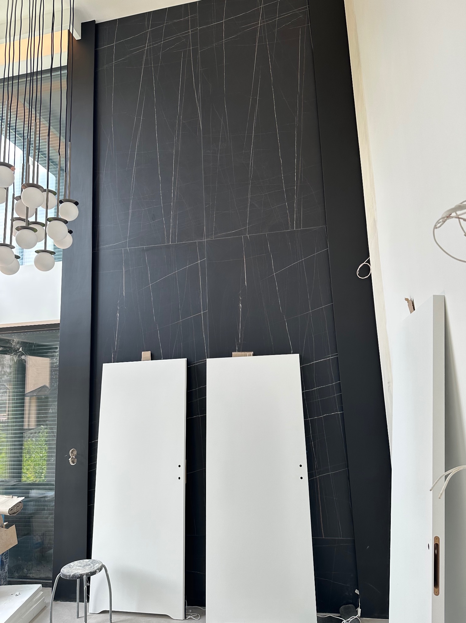 Częściowo wykończone wnętrze z czarną ścianą imitującą marmur, dwie białe, surowe płyty drzwiowe oparte o ścianę, widoczny fragment nowoczesnej lampy wiszącej i stołek roboczy.