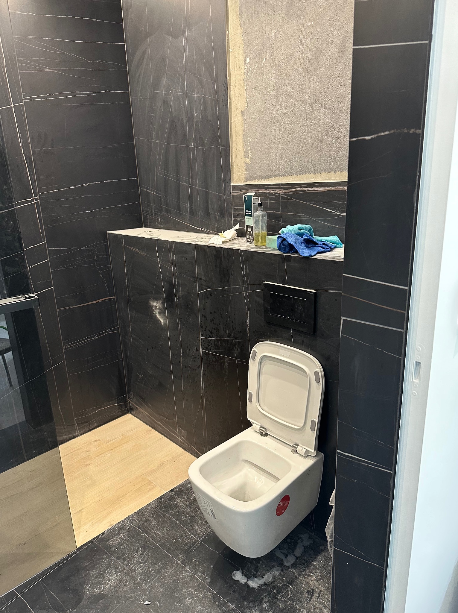 Częściowo wykończona łazienka z czarnymi płytkami imitującymi marmur, wisząca toaleta z otwartą klapą, półka z kosmetykami i niebieską ścierką, fragment surowej ściany.