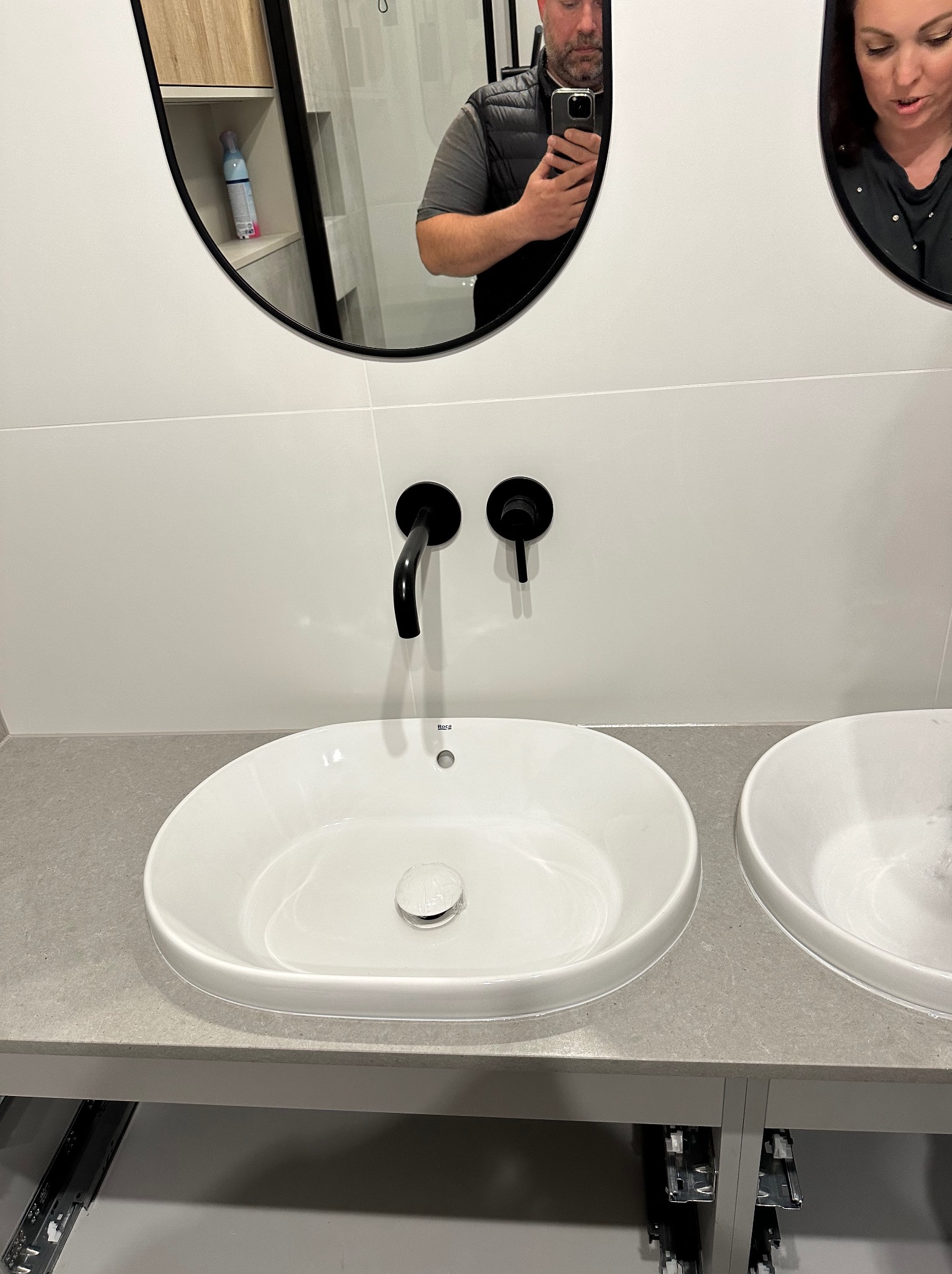 Dwie białe umywalki nablatowe z czarną armaturą zamontowane na szarym blacie, widoczne elementy montażowe szafki podumywalkowej oraz odbicie dwóch osób w lustrach nad umywalkami.