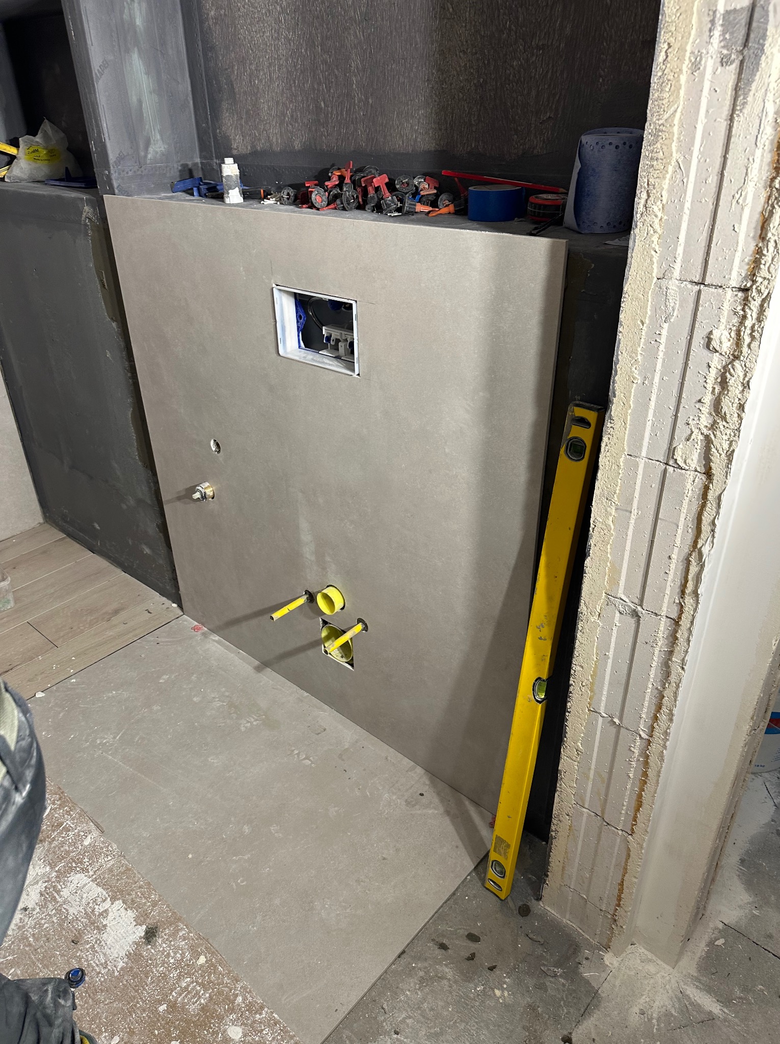 Przygotowanie ściany w łazience do montażu armatury: widoczne wyprowadzenia instalacji wodnej, otwór na stelaż WC, poziomica przy futrynie, częściowo położona podłoga z płytek.