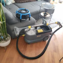 Profesjonalny odkurzacz piorący Kärcher Professional na drewnianej podłodze, obok szarej sofy z poduszkami i małego stolika, z niebieskim wentylatorem wspomagającym suszenie.