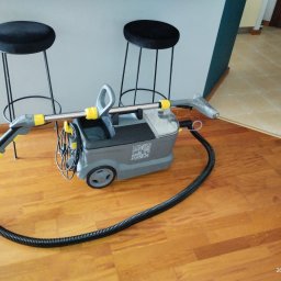 Wynajem odkurzaczy piorących - Profesjonalny odkurzacz piorący Karcher na drewnianej podłodze, z widocznymi dwoma czarnymi stołkami barowymi w tle, gotowy do użycia.