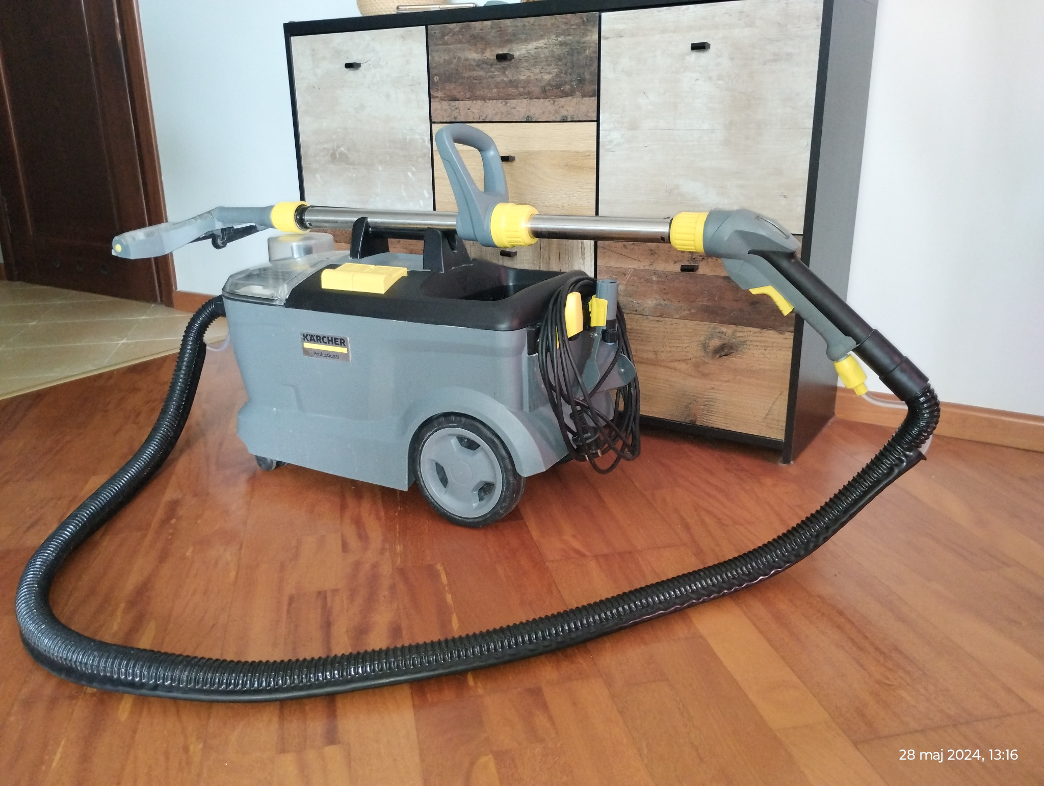 Profesjonalny odkurzacz piorący Kärcher na kółkach z długim wężem i dyszą, stojący na drewnianej podłodze, z szafką w tle.